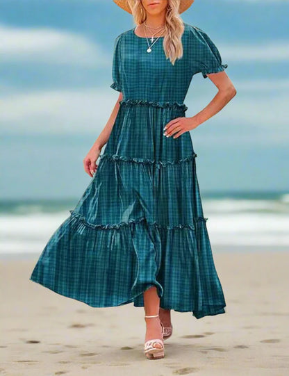 Woman wearing a teal plaid dress standing outdoors with a stone building and plants in the background.